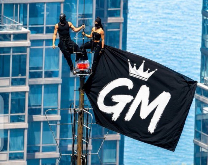 Two masked individuals stand on a pole waving a black flag with "GM" and a crown logo, set against a backdrop of modern glass skyscrapers.