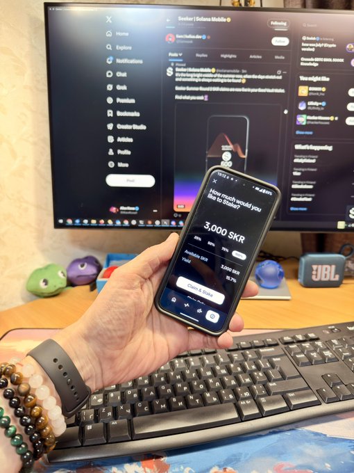 A hand holds a smartphone displaying "3,000 SKR" staked, with a monitor in the background showing the @Seeker2 Solana Mobile profile, capturing the moment of claiming the Seeker Summer reward.