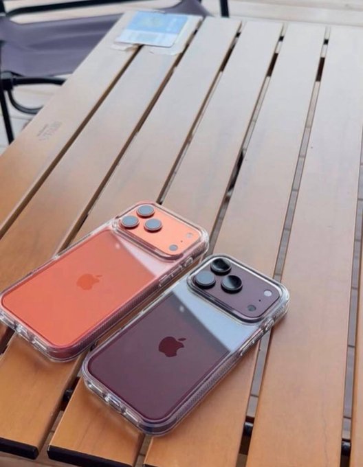 Two iPhones, one in coral orange and the other in a deep purple, rest on a wooden slatted table in clear protective cases, promoting an Apple giveaway contest.