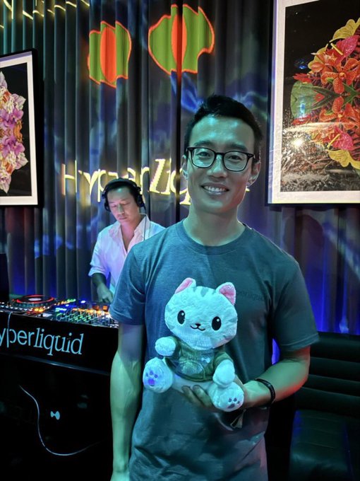 A smiling man poses holding a plush cat toy at what appears to be a Hyperliquid event, with the "HyperLiquid" branding visible on the DJ equipment and signage behind him, while a DJ performs on stage—visually reinforcing his "all in on Hyperliquid" enthusiasm.