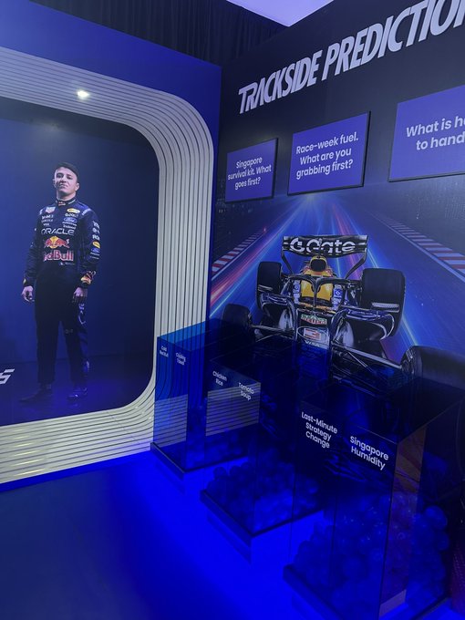 A Formula 1 exhibit displays a life-sized image of a Red Bull Racing driver in his racing suit alongside an F1 car model, with signs asking 'Trackside Predictions' questions like 'Singapore arrival. What gear first?' and 'Race-week fuel. What are you grabbing first?'
