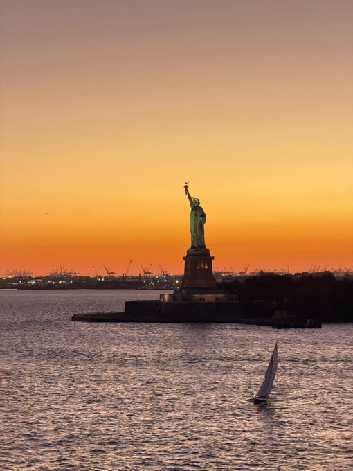 The Statue of Liberty stands against a vibrant orange sunset sky, with a small white sailboat gliding across the dark waters in the foreground.