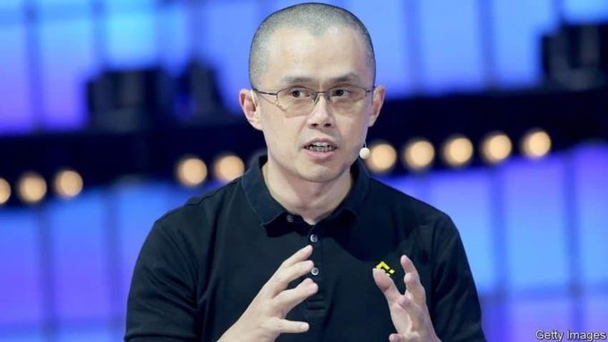 A Getty Images photo of a man with glasses and short hair wearing a black polo shirt with a yellow Binance logo, gesturing with both hands while speaking on a stage with a blue background.
