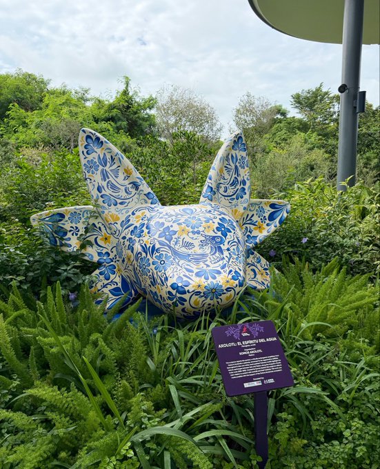 The image features an art piece, a sculpture or statue of a fish, which has blue and white colors and is surrounded by green plants. The artwork is located within a garden setting with trees and bushes around it. There are also some text elements in the image that read "From being showcased at breakpoint @somos_axolotl @SCUMSOL @solana blockchain ecosystem". This suggests that the fish sculpture was created as part of an art project or exhibition, possibly by a company like @solana, and is now displayed for visitors to enjoy.