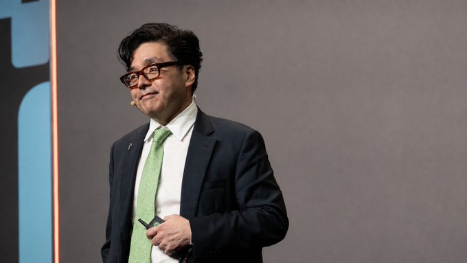A man wearing glasses, a dark suit, and a green tie stands on a stage with a gray backdrop, holding a small device in his hand.