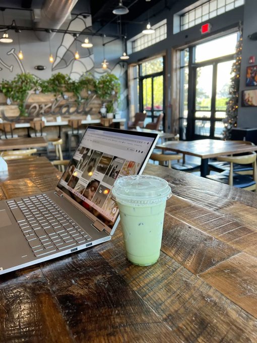 A laptop displaying charts sits open on a rustic wooden table alongside a green iced matcha latte inside a modern café with industrial décor and lush greenery. The setup perfectly captures the "gm" (good morning) post's vibe of starting the day by checking the markets from a cozy coffee spot.