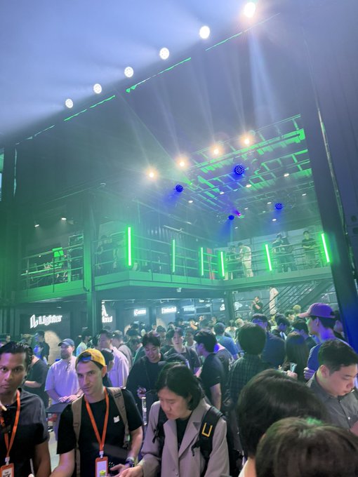 A crowded event venue illuminated with vibrant green lighting and stage lights, featuring a two-level industrial structure with people on the upper balcony and a dense crowd below.