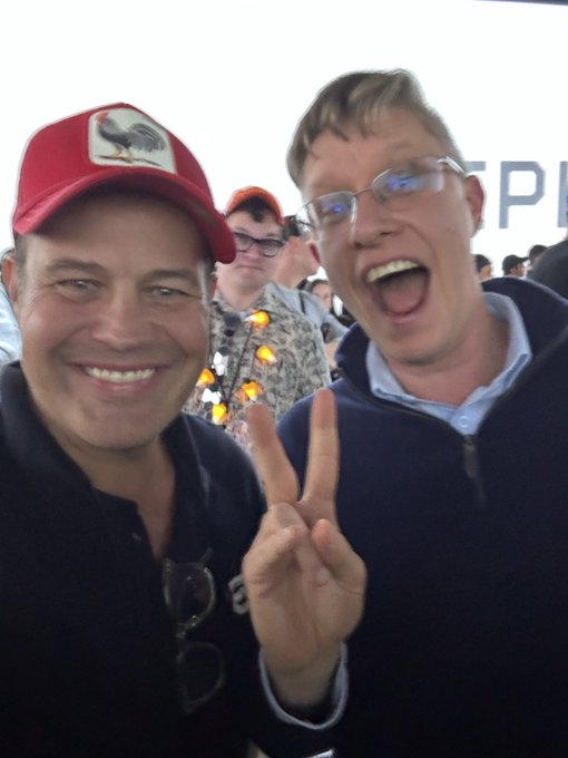Two men pose for a selfie, with the man on the left wearing a red cap featuring a rooster logo and the man on the right making a peace sign with an excited, open-mouthed expression. The image captures a lively, celebratory moment between the two, likely at an event where the bearded man on the left is "Beeple," the famous digital artist known for his "Everydays" NFT series, discussing his favorite pieces from the iconic collection.
