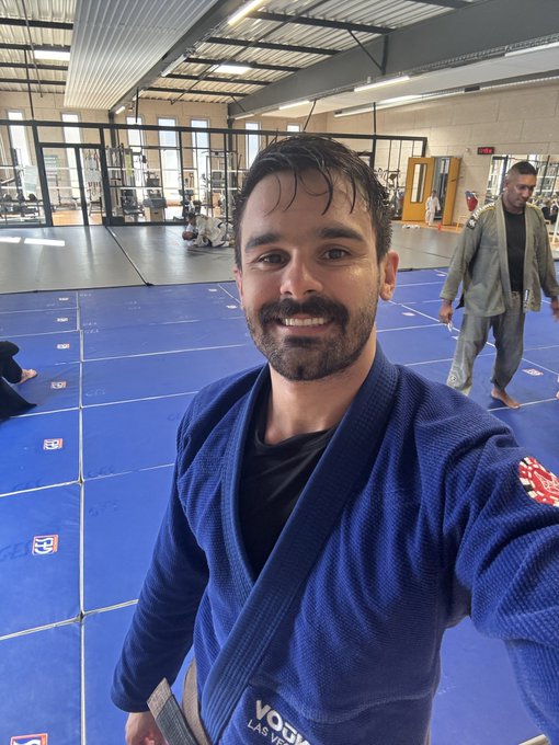 A smiling man with a mustache takes a selfie while wearing a blue Brazilian jiu-jitsu gi inside a training facility with blue mats.