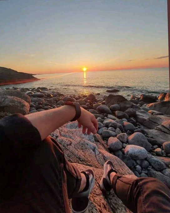 The image shows a person sitting on rocks near the ocean, looking at the sunset with their feet up. They are wearing black pants and shoes. There is also a caption that reads "Happy New Day gm At the same time, @wallchain is redefining how value is measured. Mindshare is not about the loudest voice or the highest volume. It is about consistently contributing meaningful insights that strengthen the ecosystem over time." This caption seems to be related to the image of the person sitting on rocks near the ocean at sunset and their shoes being up, which could symbolize a moment of relaxation or contemplation during an otherwise busy day.