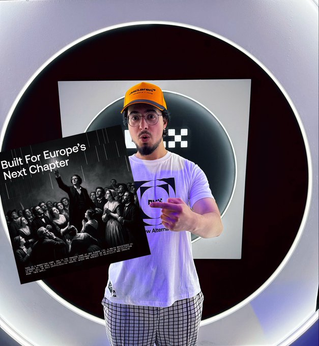 A man wearing a McLaren orange cap and a white t-shirt stands in a circular illuminated setting, holding up a placard reading "Built For Europe's Next Chapter" featuring an artistic rendering of a crowd scene, visually echoing the post's message about MiCA compliance and @OKX's regulated status ahead of the July 1 deadline.