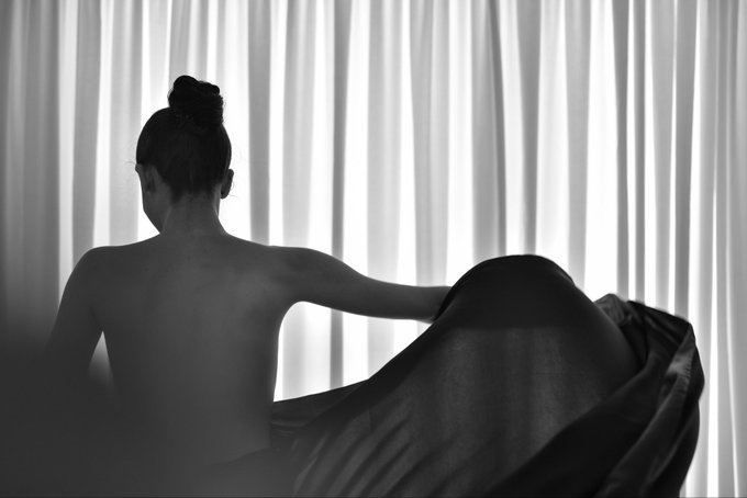 A black and white silhouette photograph showing a woman with her hair in a bun, sitting with her back to the camera in front of sheer curtains, holding the fabric to one side.