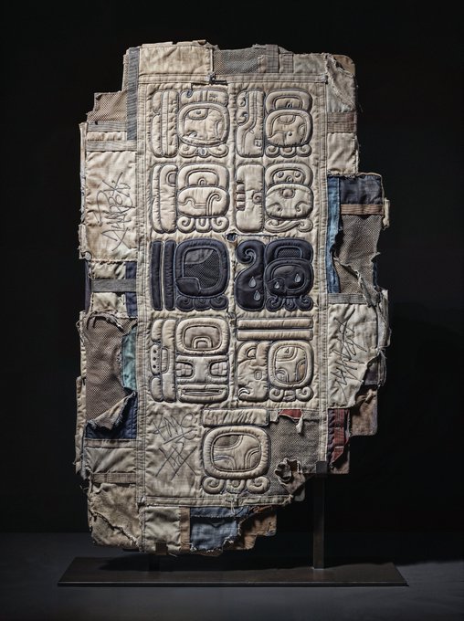 An ancient Maya carved stone stela featuring stacked glyphs and a central figure, displayed upright on a black museum mount against a dark background.