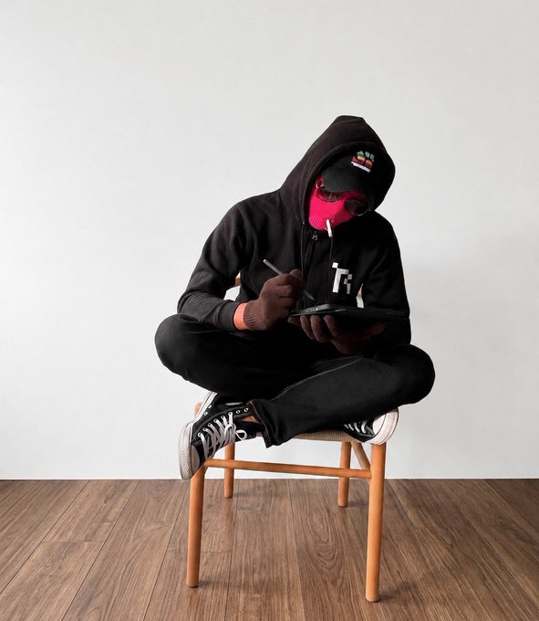 A person dressed entirely in black with a hood, cap, and pink face covering sits cross-legged on a wooden stool, writing or drawing on a tablet in a minimalist room with white walls.