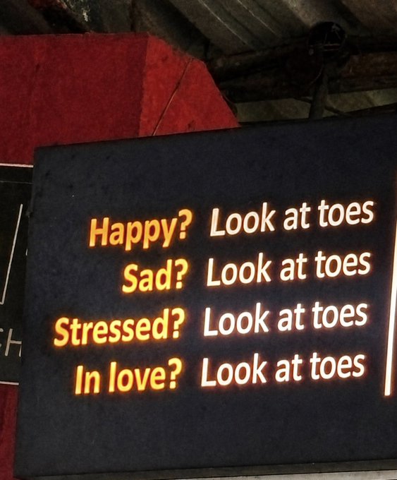 A backlit sign humorously advises "Happy? Sad? Stressed? In love? Look at toes," which ties in perfectly with the accompanying "$TOESCOIN" post by promoting an all-encompassing focus on toes regardless of one's emotional state.