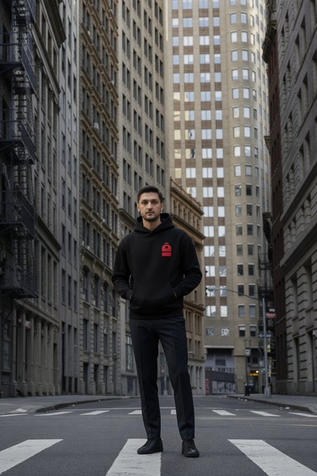 A man wearing a black hoodie with a red logo and dark pants stands at a crosswalk on an empty city street lined with tall buildings.