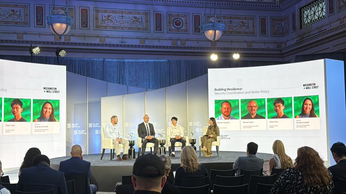 A panel discussion taking place on a stage at a Wall Street event, featuring five speakers seated in chairs with large screens behind them displaying 'Building Resilience: Security Coordination and Better Policy' and speaker headshots.