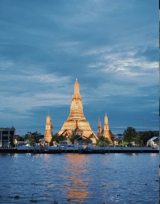 A large, ornate temple complex with a central spire and smaller surrounding towers, illuminated in warm light against a dusky blue sky, is reflected in a body of water in the foreground.