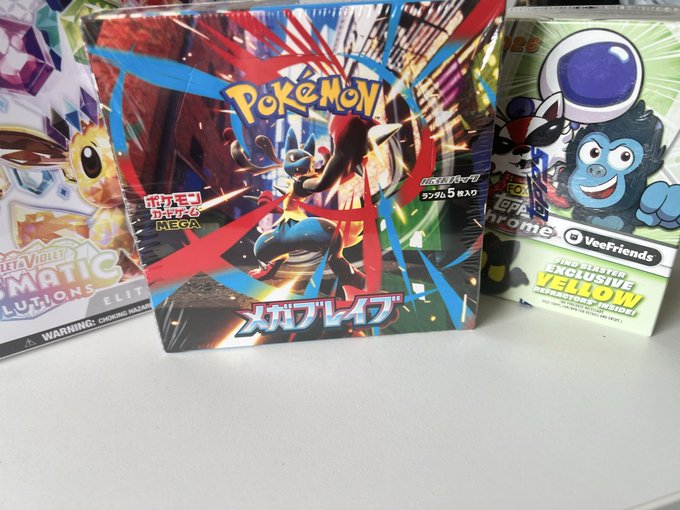 A photograph of Pokemon trading card game products, including a Japanese 'Mega Brave' booster box in the center flanked by Sleevmatic Elite box on the left and an Exclusive Yellow Veefriends box on the right.