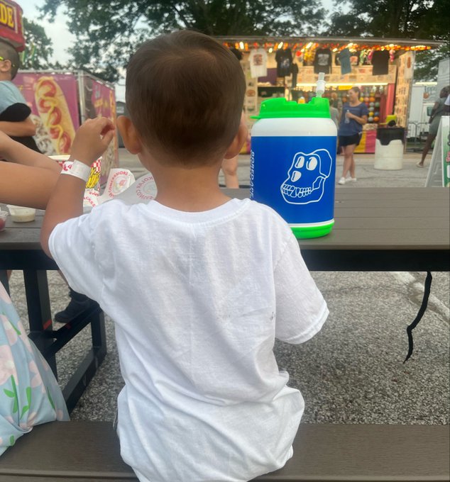 A young boy in a white t-shirt sitting at a wooden picnic table, facing away from the camera, with a large blue and green water bottle featuring a skull design on the table in front of him at what appears to be a fair.