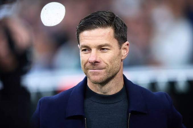 A close-up portrait of a man in a dark blue coat over a black shirt, smiling slightly with a blurred stadium background.