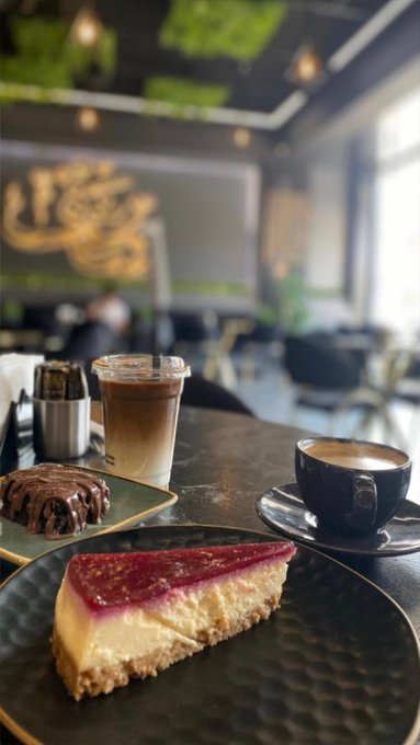 A photo of a dessert spread on a dark table in a cafe, featuring a slice of cheesecake with red topping, a chocolate pastry, an iced latte in a plastic cup, and a cappuccino in a black cup.