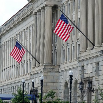 Two American flags hang from the ornate stone facade of a grand classical building featuring tall columns and arched windows.