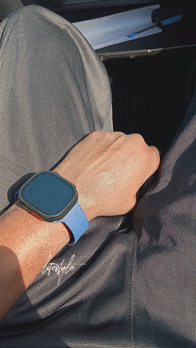 A person's wrist wearing an Apple Watch with a blue band rests on their lap, with a signature visible in the corner of the image.