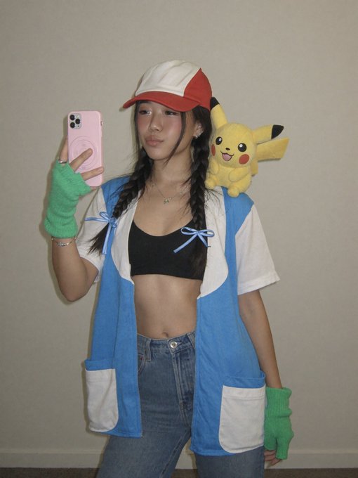 A young woman in an Ash Ketchum-inspired costume with a red and white cap, blue vest, and green fingerless gloves, taking a mirror selfie with a pink phone while a Pikachu plush sits on her shoulder.