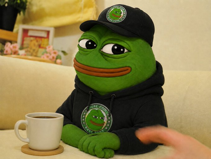 A Pepe the Frog plush toy wearing a 'PEPE COIN' cap and hoodie, sitting on a couch next to a coffee mug.