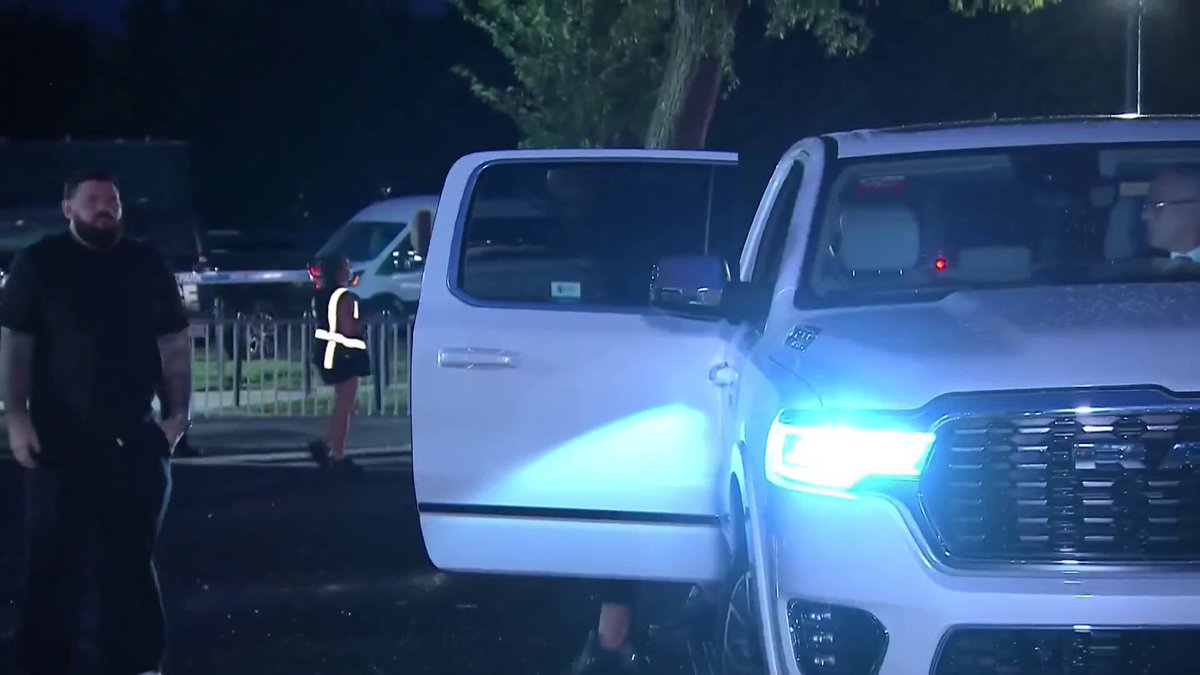 The image shows a white truck parked next to a fence, with two people standing near the door of the truck. The scene appears to be at a press event for UFC, as suggested by the text "UFC White House". One person seems to be wearing a black shirt and jeans, while another is dressed in a white shirt and blue jeans. A car can also be seen parked nearby.