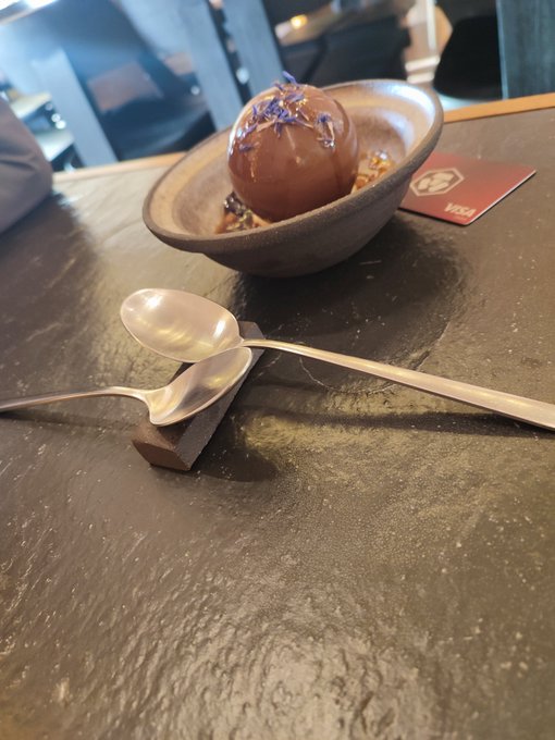 A chocolate sphere dessert garnished with edible flowers sits in a brown ceramic bowl on a dark stone table, accompanied by two silver spoons on a rest and a reserved placard.