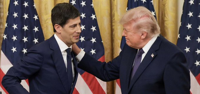 Two men in dark suits stand in front of American flags, with one placing his hand on the other's shoulder.