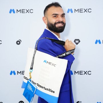 A smiling man, identified in the post as the CEO, poses at a MEXC event while holding a MEXC badge on a lanyard and touching his suit jacket. The backdrop repeatedly displays the “MEXC” logo, and his badge reads “CEO” and “Global Head Opportunities,” matching the post’s playful comment about a favorite side quest.