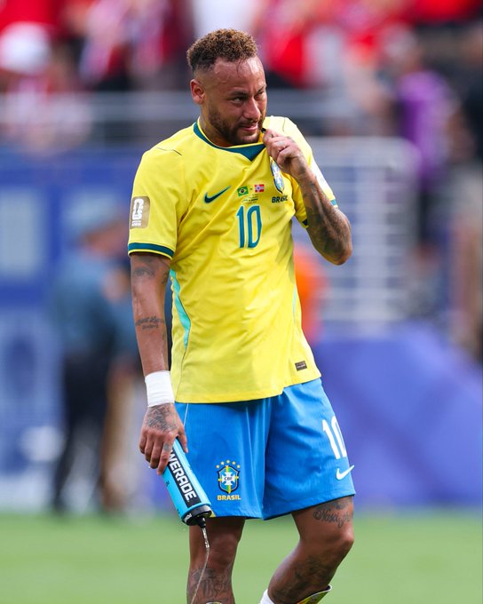 Brazilian soccer player Neymar, wearing the #10 yellow Brazil national team jersey and blue shorts, walks on the field while drinking from a Powerade bottle.