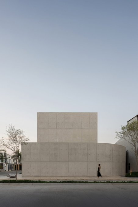 A minimalist concrete building stands prominently against a clear sky, with a solitary figure walking past its smooth, geometric facade. The architectural simplicity and contemplative atmosphere evoke the serene, meditative aesthetic often associated with the Baha'i faith, subtly complementing the post's poetic notion of $TAO one day enabling the purchase of "Casa Tao."