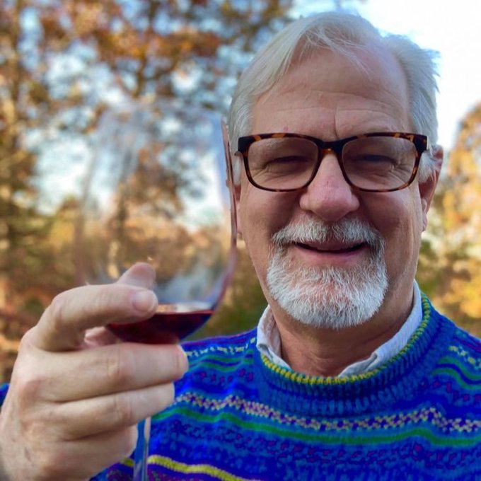 An older smiling man with white hair and beard, wearing tortoiseshell glasses and a blue patterned sweater, holding up a glass of red wine outdoors.