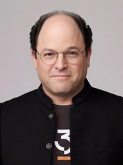 A headshot photo of a balding man with glasses wearing a black jacket over a t-shirt with a white graphic design, posing against a gray background.