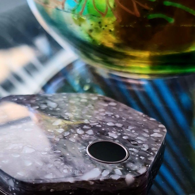 A close-up shot of a mottled brown and white sink with water droplets, featuring a dark circular drain stopper and a glass vessel with green and yellow liquid behind it.