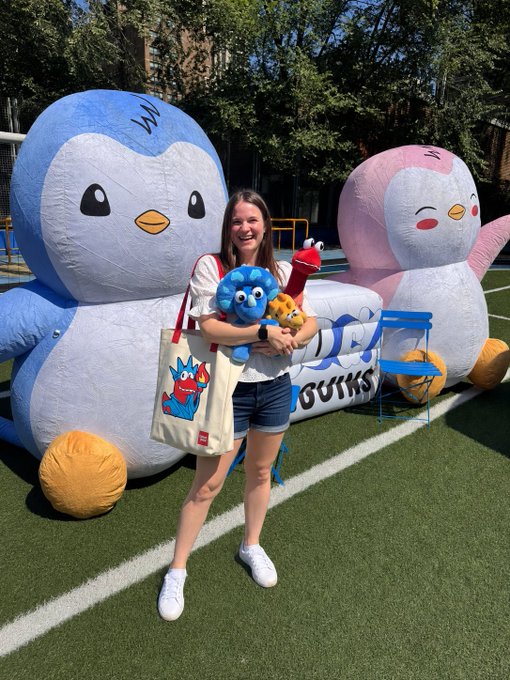 A smiling woman on a sports field standing between large inflatable penguin mascots (one blue, one pink), holding plush toys and wearing a tote bag with a cartoon character design.