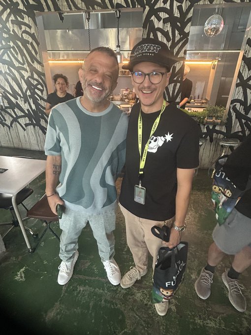 The image features two men standing next to each other, both wearing glasses and smiling for a photo. One of them has a black bag with white text on it. The background includes a dining table and chairs, suggesting that they might be at an event or gathering. There is also another person visible in the scene, but their face is not clearly visible due to the focus on the two men posing together.