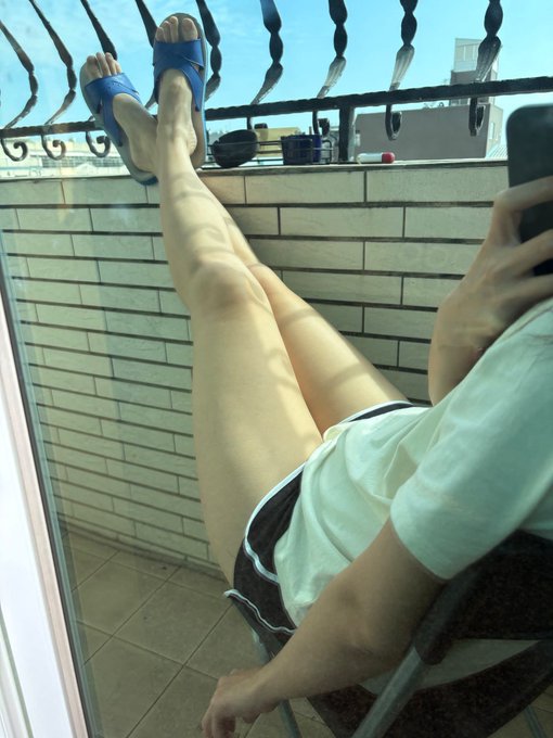 A person takes a mirror selfie on a balcony, wearing white shorts and blue sandals with their legs propped up against the railing, relaxed under a sunny sky. The leisurely pose visually contrasts with the post's energetic demand for Bitcoin to reach 100,000 ("BTC 100kkkkkkk"), playfully suggesting they're chilling while waiting for that price target.