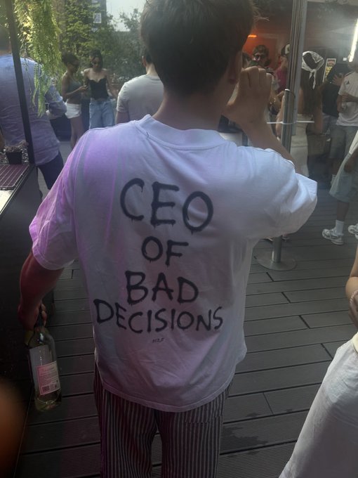 A person seen from behind wearing a white t-shirt with the handwritten text 'CEO OF BAD DECISIONS' holds a bottle at what appears to be an outdoor social gathering.