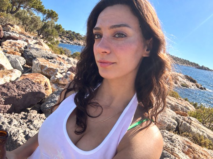 A young woman with long dark wavy hair wearing a white halter top poses for a selfie on rocky terrain by the sea.