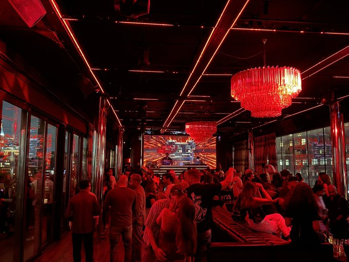 A crowded nightclub or bar interior bathed in red lighting with geometric LED ceiling strips, ornate chandeliers, and a large screen displaying neon-style graphics.