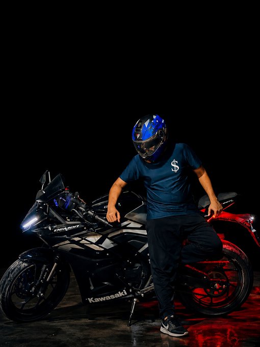A person in a blue shirt, dark pants, and a blue helmet poses next to a black Kawasaki motorcycle in a dimly lit setting with red lighting.