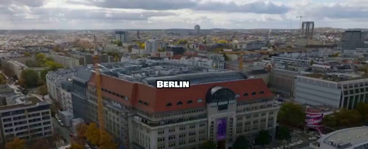 The image shows an aerial view of a city with a large building featuring the word "Berlin" written on its roof. There are also other buildings visible, indicating that this is a bustling urban area. The photo appears to be taken during the day under cloudy skies, suggesting it was captured in the middle of the afternoon or early evening hours.