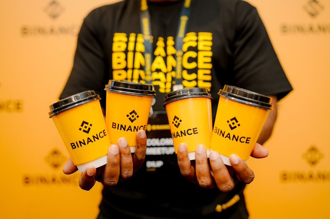 A person wearing a Binance-branded shirt holds four yellow Binance coffee cups against a Binance-logoed backdrop. The image pairs with the "GM☕️ Monday wake-up" post, visually riffing on the idea of "good morning" coffee while nodding to the crypto exchange's brand.