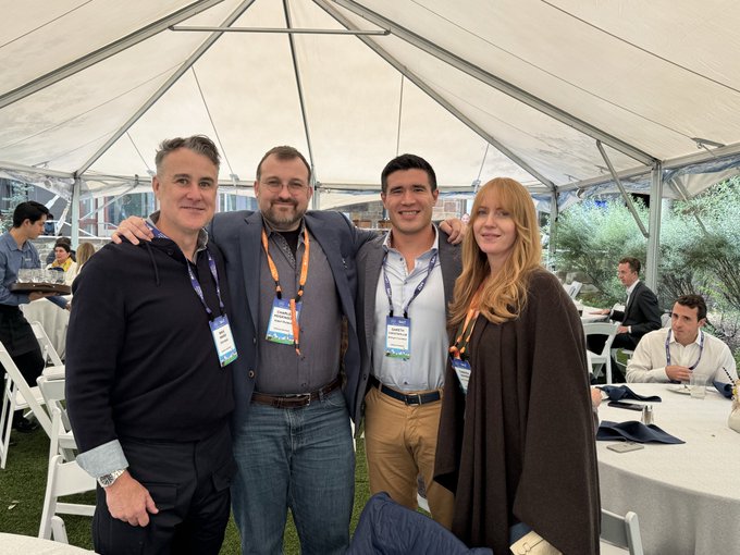 Four people pose together for a photo at an outdoor event under a tent, all wearing conference lanyards with badges.
