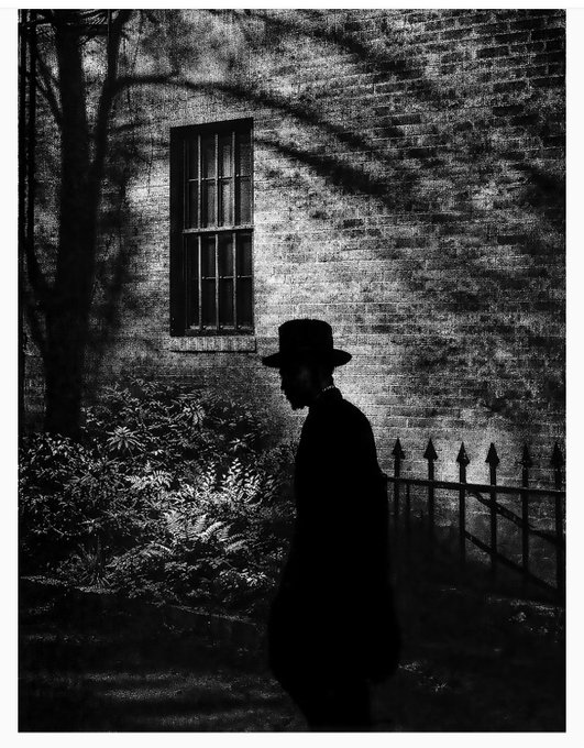A black and white artistic photograph of a silhouetted figure wearing a wide-brimmed hat walking past a brick building with a barred window and wrought iron fence.